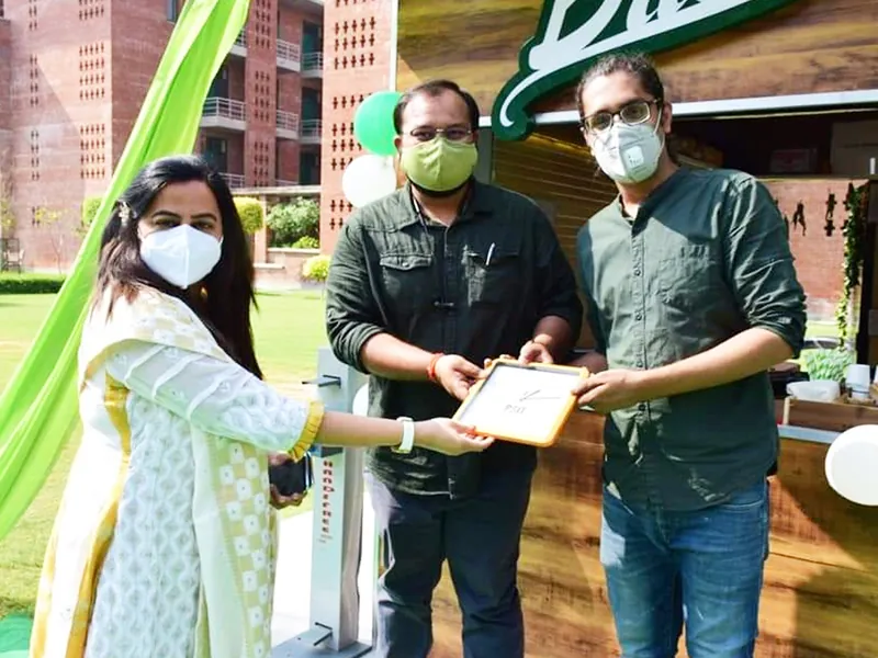 Three people in face masks hold a framed plaque marked PSIT in front of a BRIO kiosk